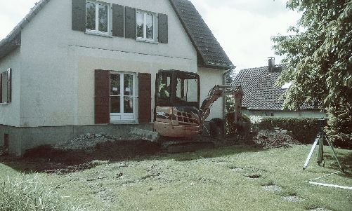 Maison blanche avec des volets rouges et une terrasse en bois dans un jardin gazonné, avec un grand arbre sur la droite.