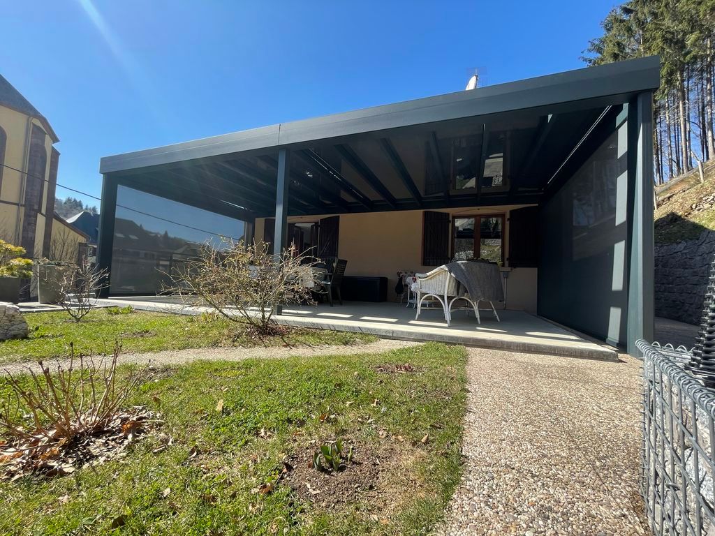 Patio de maison moderne avec une pergola vitrée, des sièges extérieurs et une cour avant gazonnée sous un ciel bleu.