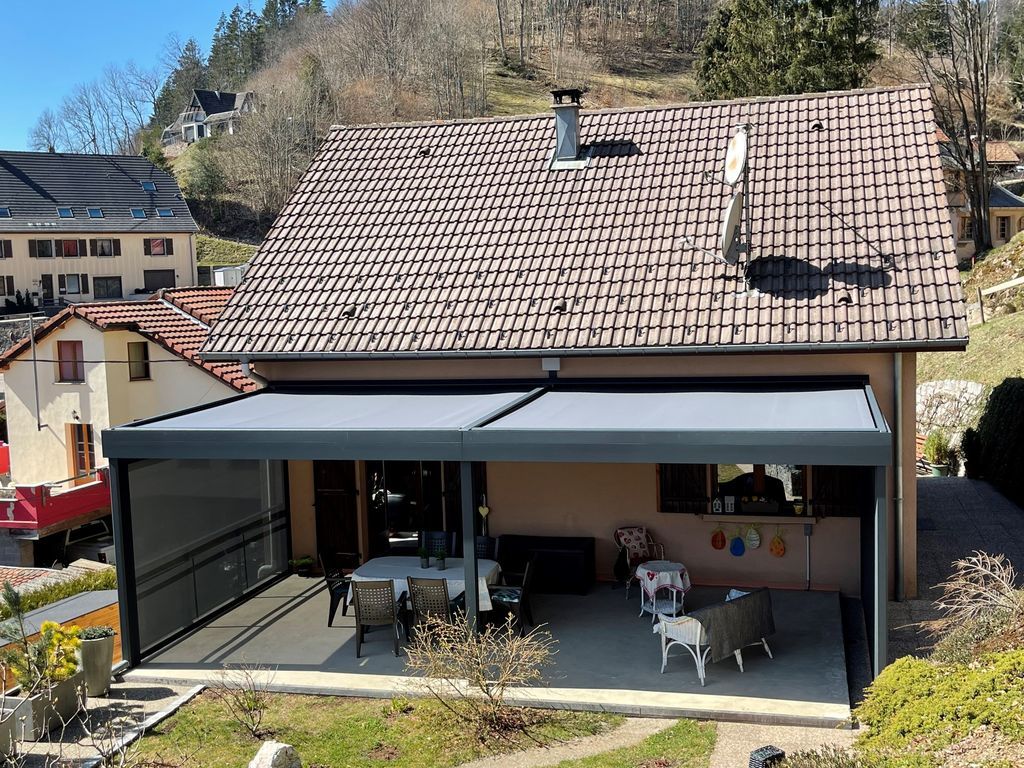 Maison avec toit de tuiles et terrasse couverte dans un village à flanc de colline, avec mobilier de jardin et petit jardin.