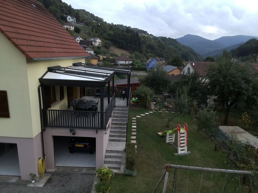 Maison au toit rouge jouxtant une cour herbeuse, surplombant un village à flanc de colline et des montagnes sous un ciel nuageux.