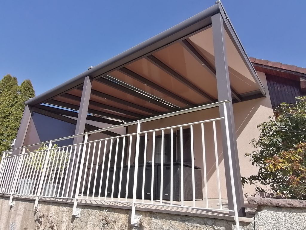 Terrasse extérieure couverte avec une structure métallique grise, des panneaux de toit beiges et une rambarde blanche, jouxtant une maison.