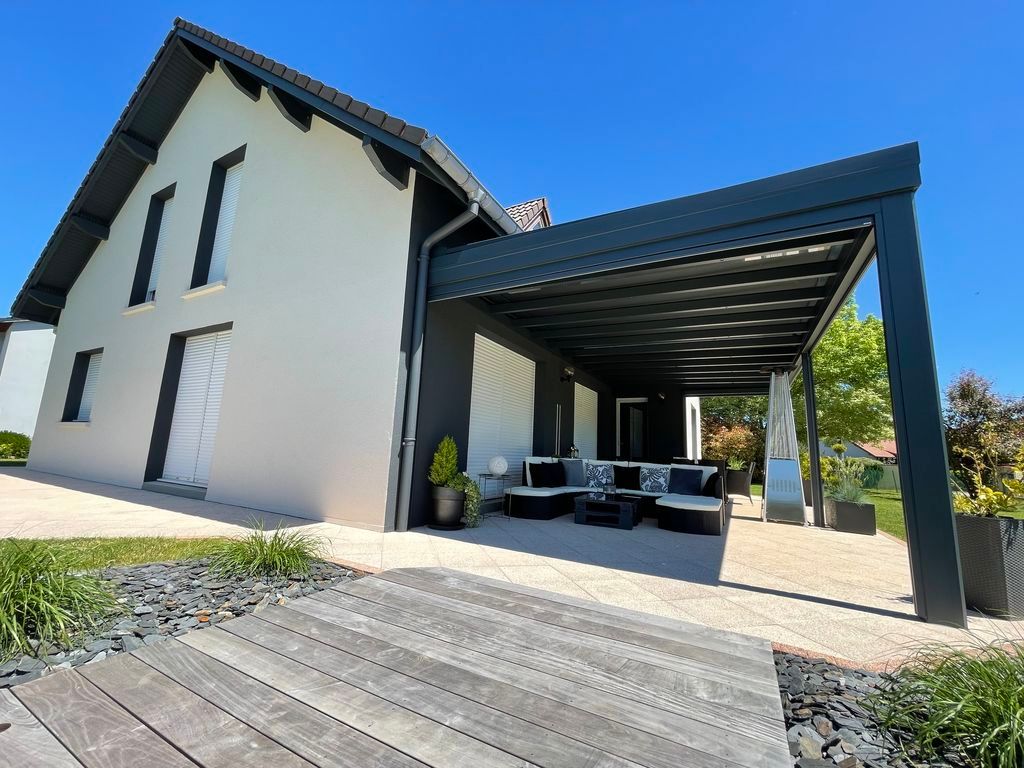 Maison moderne à deux étages avec patio couvert, coin salon extérieur et terrasse en bois sous un ciel bleu azur.