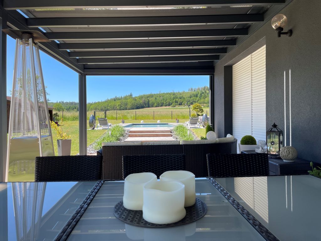 Patio moderne avec table à manger, bougies blanches et vue sur les collines verdoyantes sous une pergola couverte.