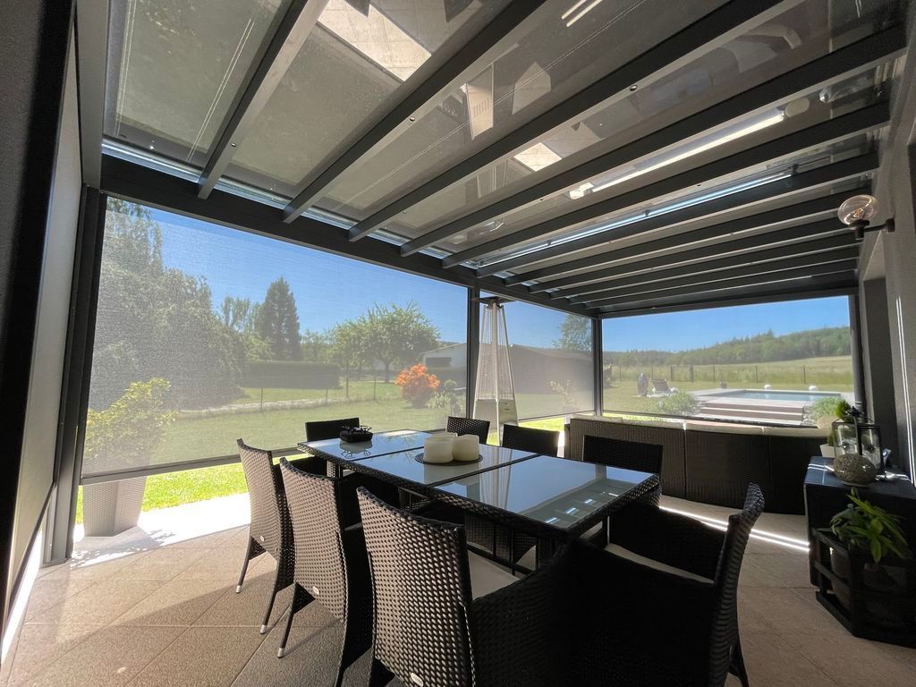 Terrasse couverte avec table à manger en verre, chaises noires et vue sur la montagne sous une pergola moderne.