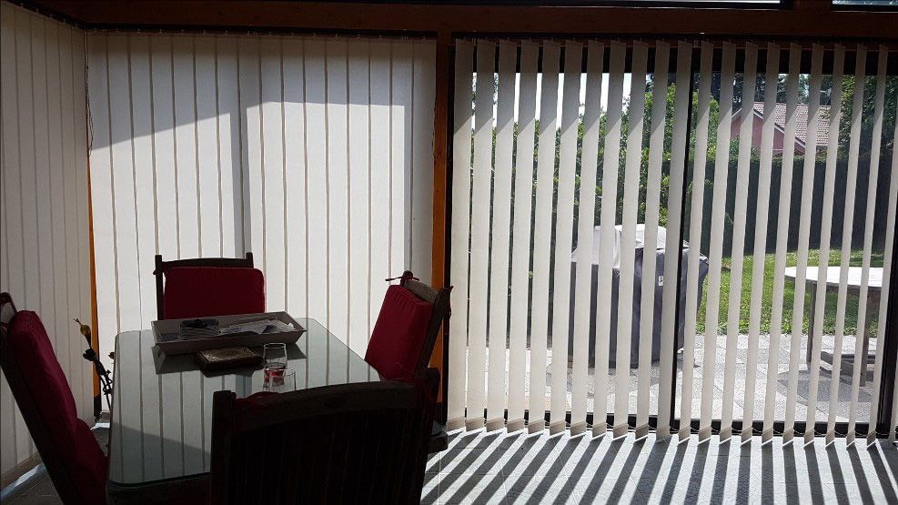 Coin repas avec table en verre et chaises rouges, à côté de stores verticaux et d'une porte-fenêtre lumineuse