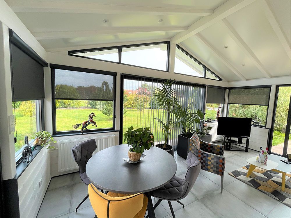 Véranda lumineuse avec table ronde, chaises, plantes d'intérieur et grandes fenêtres donnant sur un jardin verdoyant