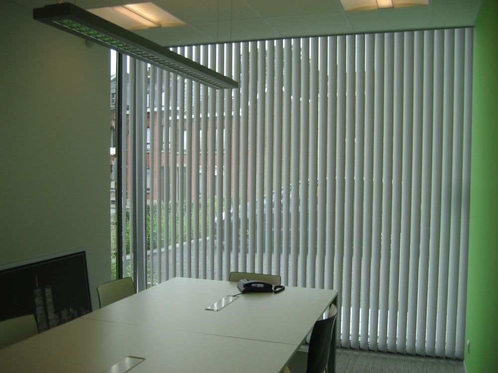 Salle de conférence vide avec murs verts, table, chaises et stores verticaux aux fenêtres.