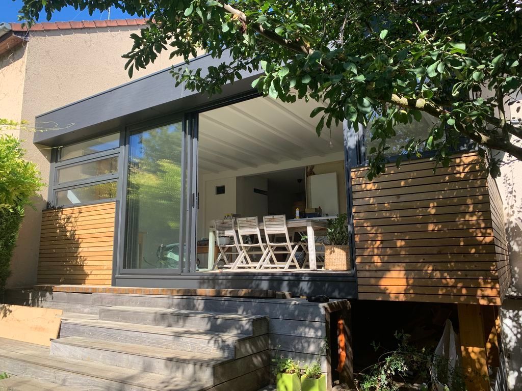 Patio de maison moderne avec portes vitrées, bardage en bois et ensemble de repas extérieur sous un arbre feuillu.
