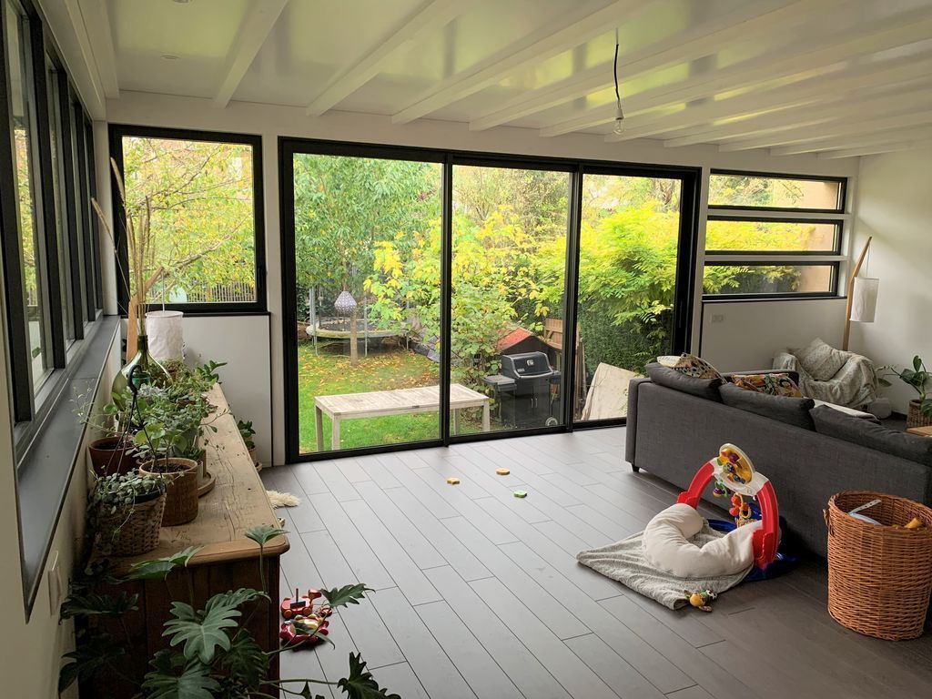 Véranda lumineuse avec portes vitrées, plantes, canapé gris et un enfant assis sur un coussin près d'un panier en osier.