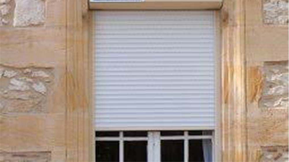 Volet blanc fermé sur un bâtiment en pierre beige, au-dessus d'une petite fenêtre.