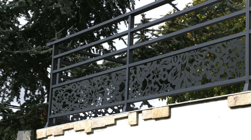 Rambarde en métal noir ornée de panneaux décoratifs au-dessus d'un mur blanc, avec des arbres en arrière-plan.