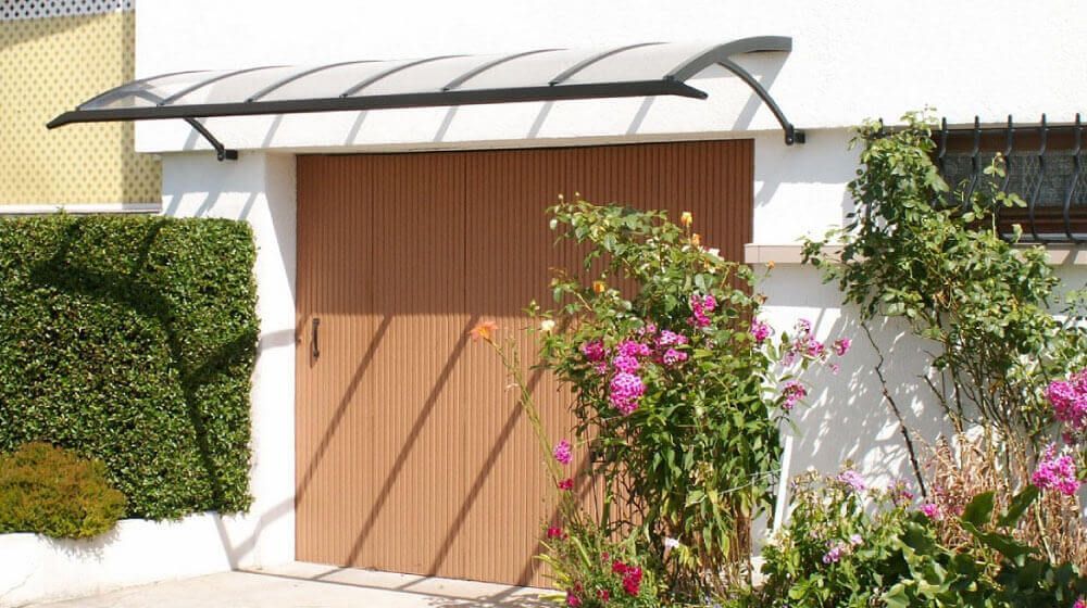 Entrée d'une maison blanche avec portail en bois, auvent métallique incurvé et arbustes fleuris devant