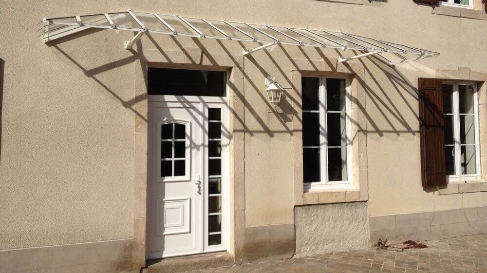 Façade de maison beige avec porte et fenêtres blanches, recouverte d'une structure métallique auvent.