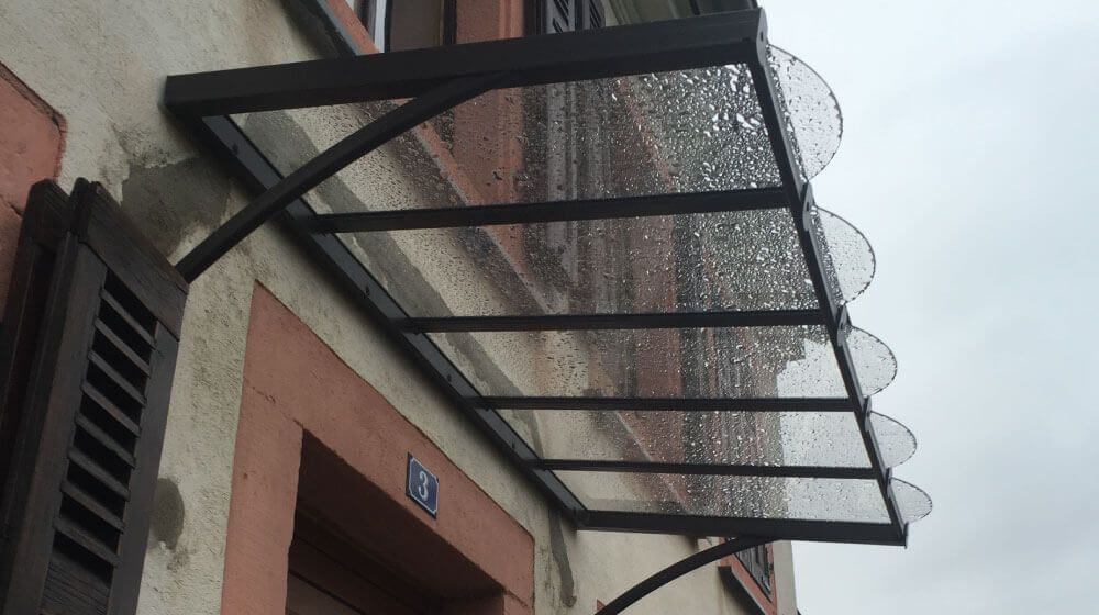 Auvent en métal noir avec panneaux arqués transparents au-dessus d'une entrée de bâtiment rose