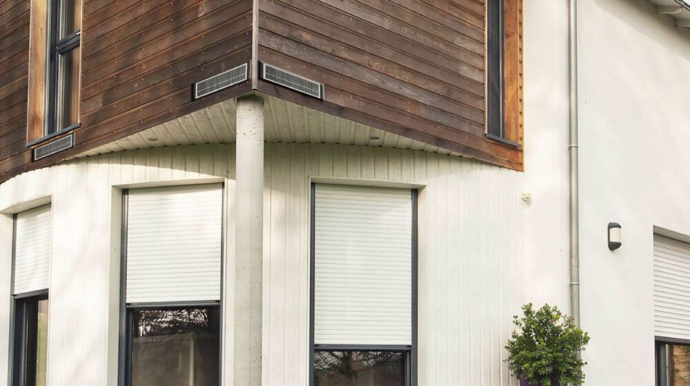 Coin d'un bâtiment moderne aux murs blancs, au bardage en bois brun et aux fenêtres et volets fermés.
