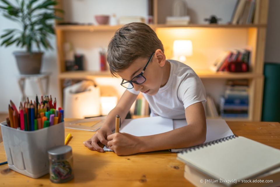 Junge mit Brille schreibt an einem Holzschreibtisch mit Buntstiften und einer Lampe in ein Notizbuch.