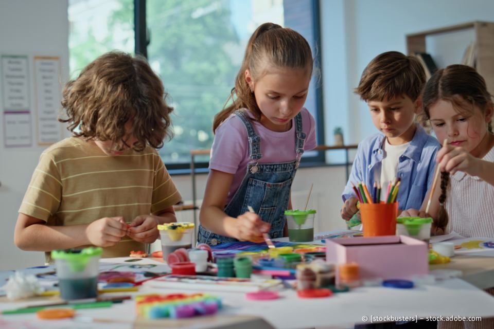 Vier Kinder malen an einem Tisch in einem Klassenzimmer; ein Mädchen mit Pferdeschwanz malt mit einem Pinsel.