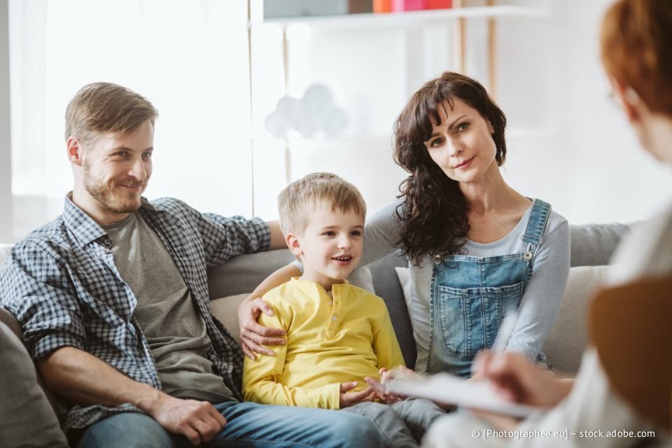 Eine dreiköpfige Familie sitzt mit einem Therapeuten zusammen. Die Eltern lächeln, das Kind wirkt neugierig; der Raum ist hell.