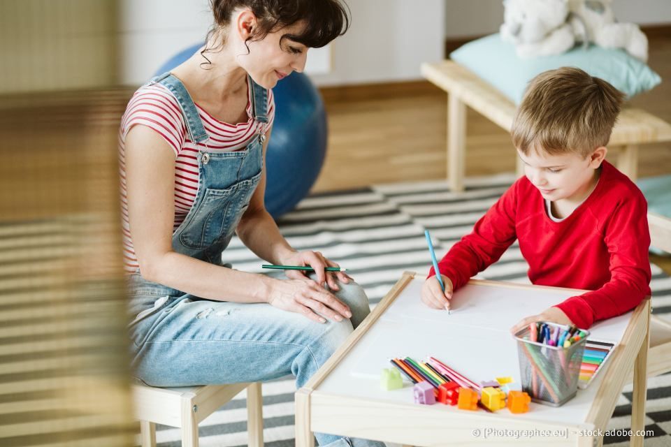 Frau und Kind malen gemeinsam an einem kleinen Tisch. Die Frau lächelt. Bunte Buntstifte sind zu sehen.