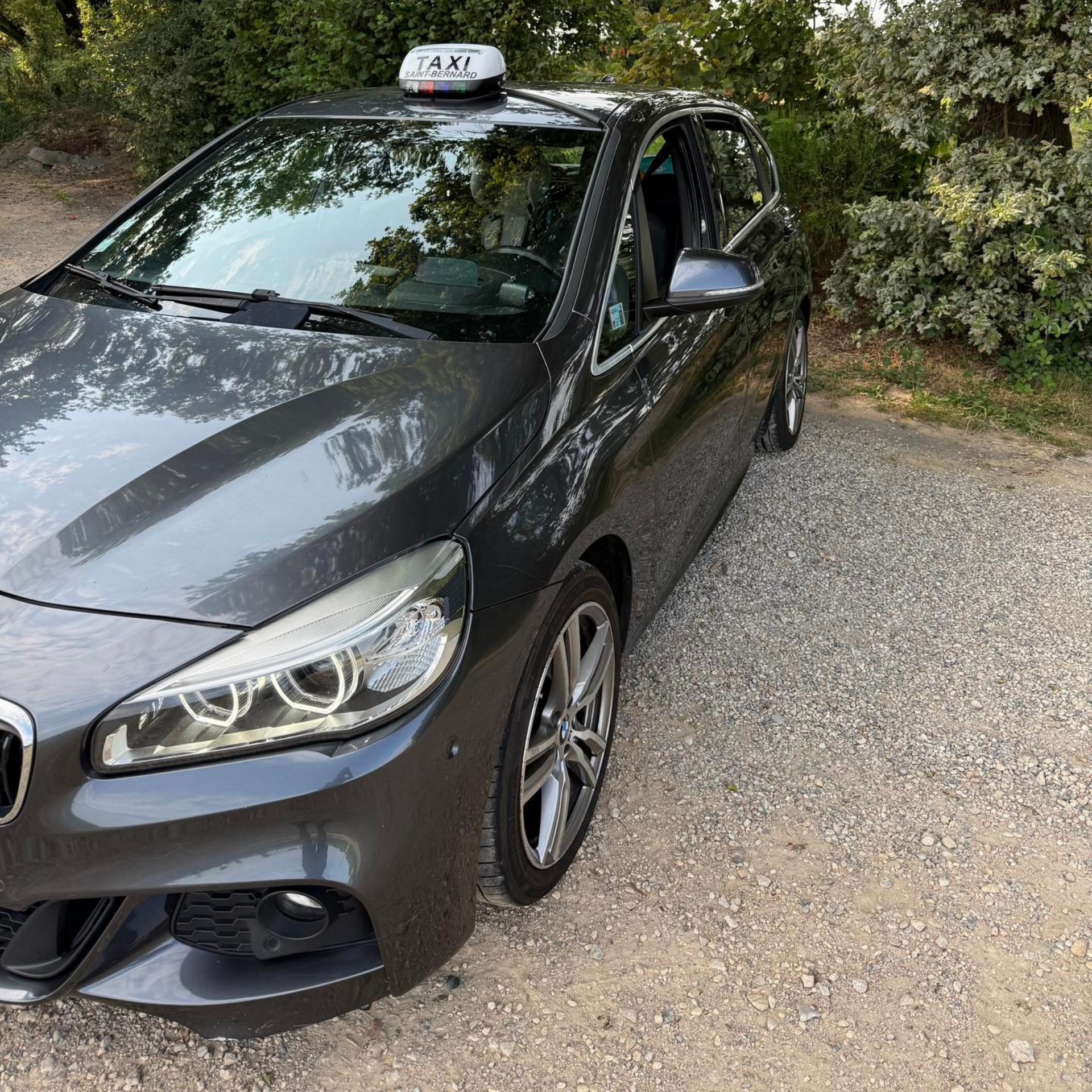 Taxi BMW gris stationné sur un chemin avec des arbres.