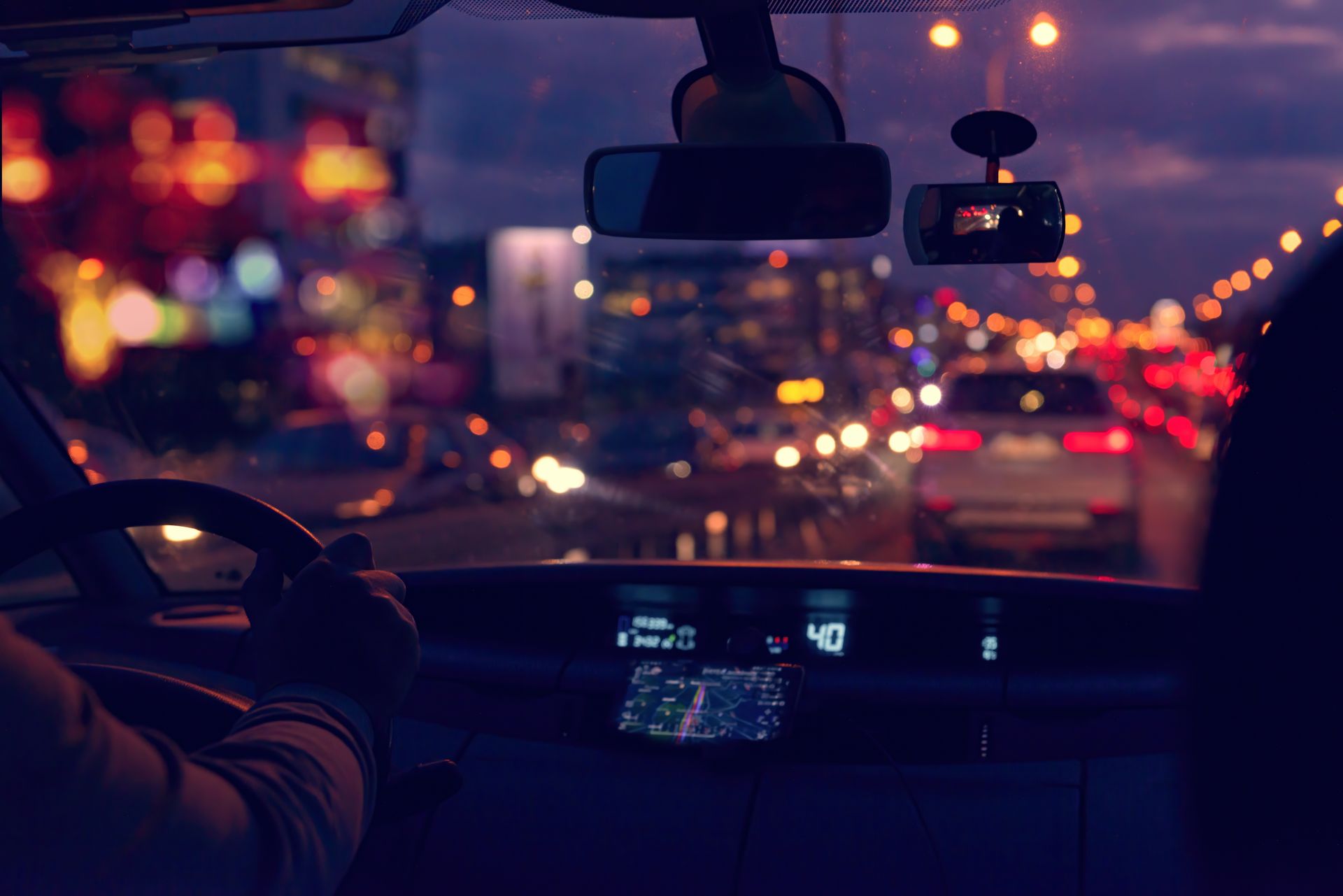 Vue depuis l'intérieur d'une voiture roulant de nuit. Les lumières de la ville se fondent en arrière-plan. Les mains du conducteur sont sur le volant.