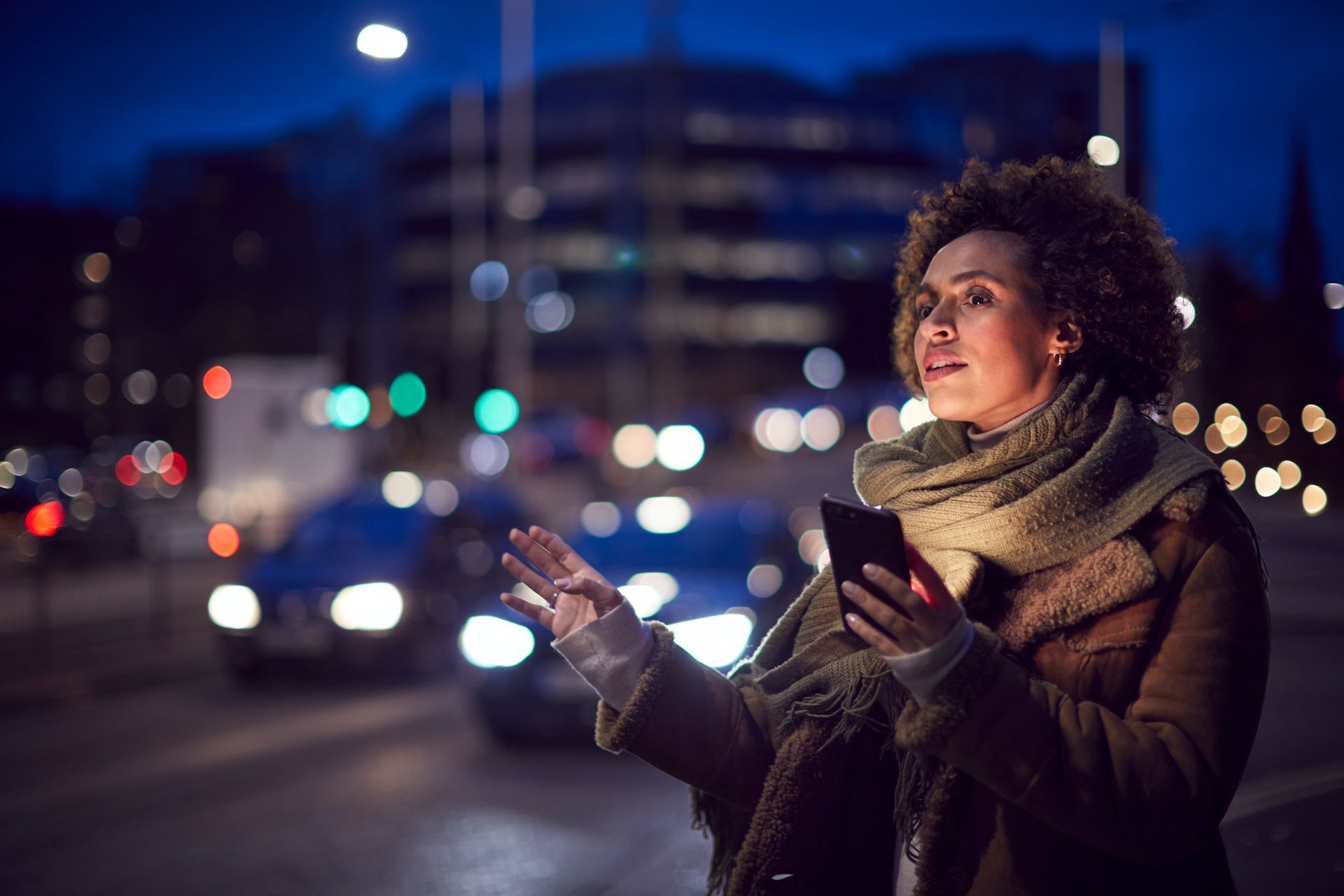 Une femme dans une rue de la ville, la nuit, hèle un taxi, son téléphone à la main.
