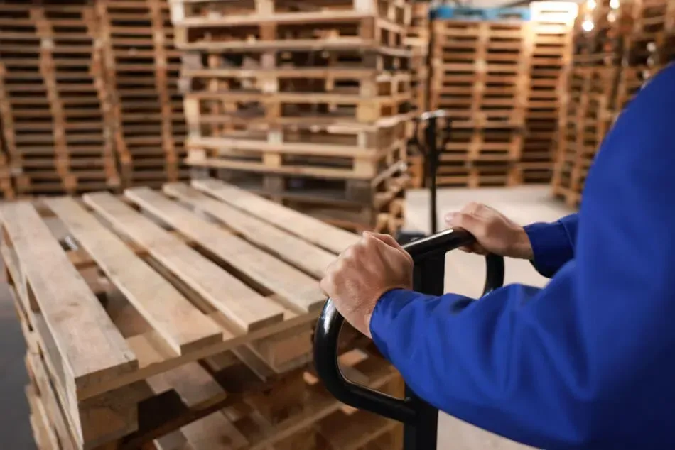 Un hombre empuja una carretilla manual con paletas de madera en un almacén.