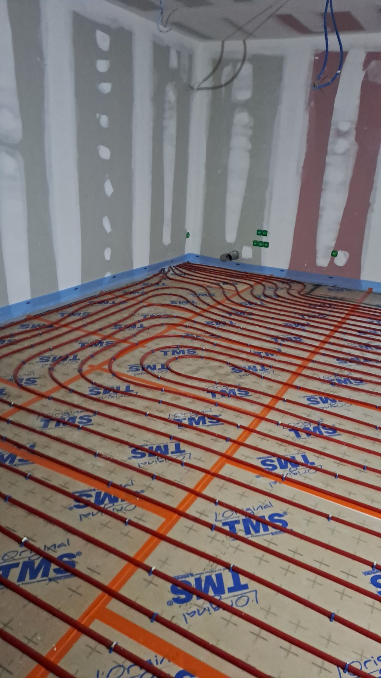 Des tuyaux de chauffage au sol radiant orange sont installés sur des panneaux isolants dans une pièce aux murs en plaques de plâtre non finies