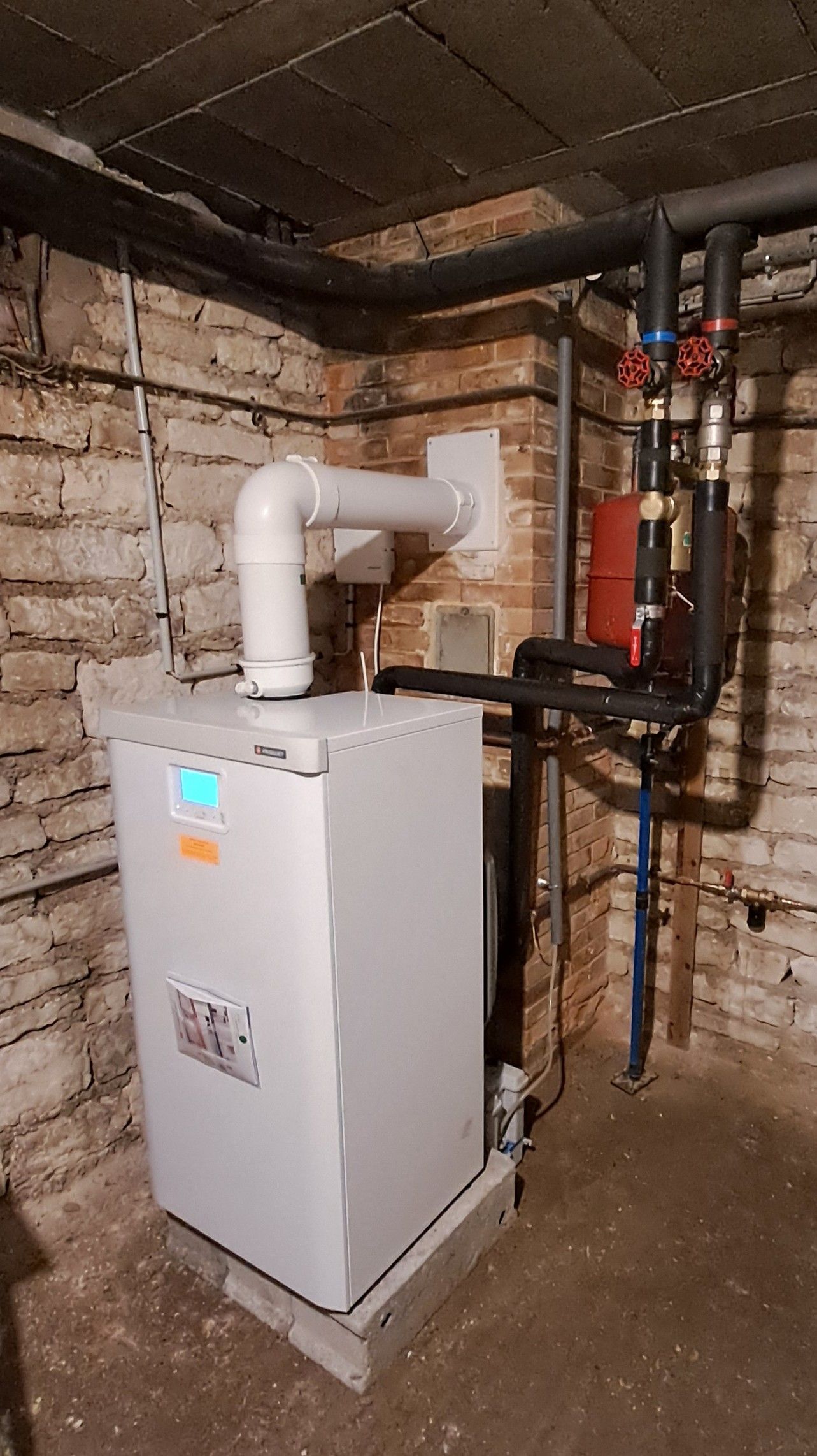 Dans un sous-sol aux murs de briques apparentes, une chaudière à gaz blanche et moderne est installée, reliée à des tuyaux et à une sortie d'évacuation blanche