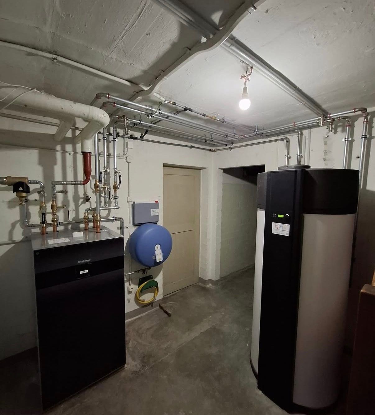 Hauswirtschaftsraum im Keller mit Heizkessel, Rohren, Wassertank und freiliegender Deckenleuchte.