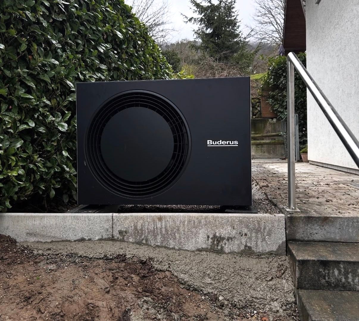 Schwarze Aussenwärmepumpe neben Betonstufen und einer weissen Wand in einem Gartenbereich