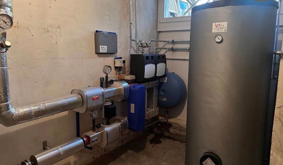 Heizraum mit Rohren, Pumpen und einem grossen Grauwasserspeicher in der Nähe eines Kellerfensters.