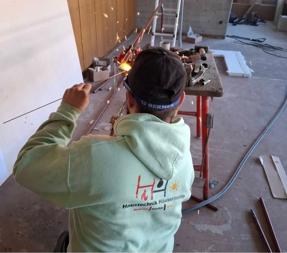 Eine Person schweisst in einer Werkstatt einen Metallrahmen, Funken sprühen, sie trägt einen grünen Kapuzenpullover und eine grüne Kappe.