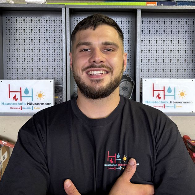 Ein lächelnder Mann in schwarzem T-Shirt steht vor Regalen mit Markenverpackungen und einem Logoschild.