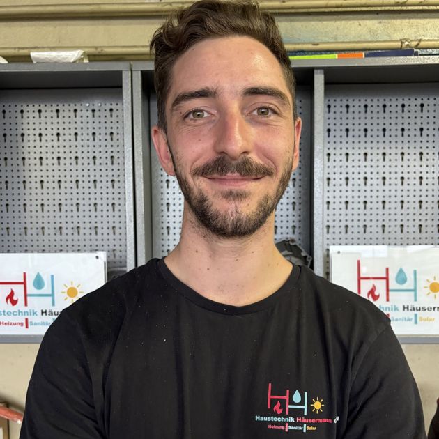 Ein Mann in einem schwarzen T-Shirt lächelt vor Büroregalen und einer Lochwand.