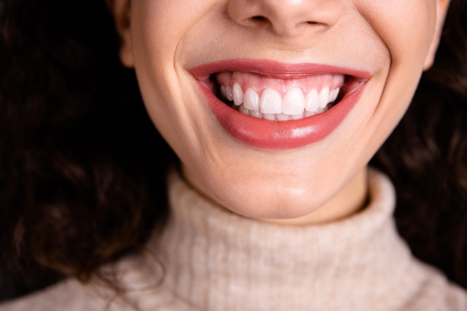 Primer plano de una boca sonriente con lápiz labial rojo brillante y dientes visibles en un suéter de cuello alto beige.