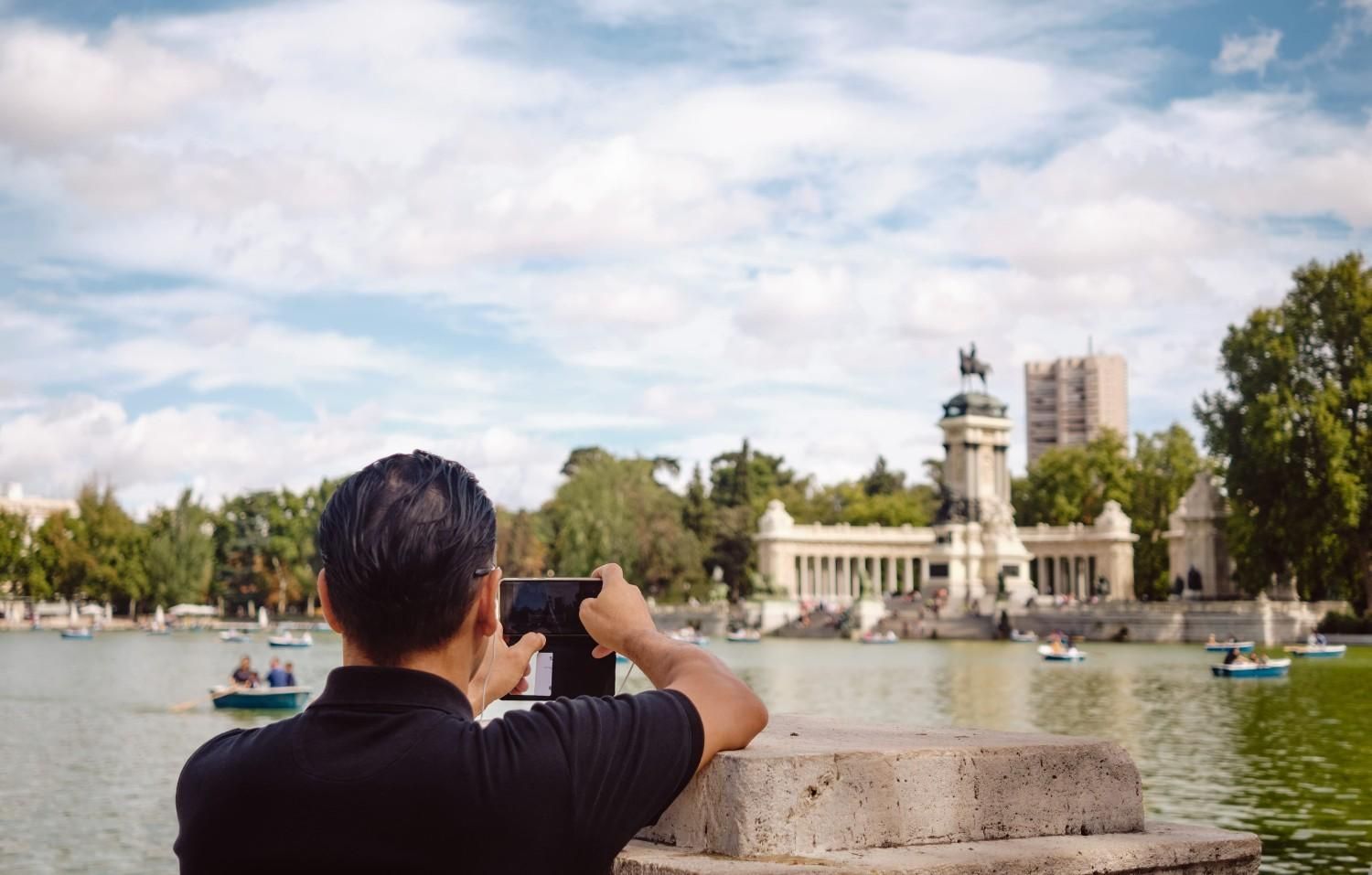 Person photographing a lakeside park with a fountain and city buildings under a cloudy sky