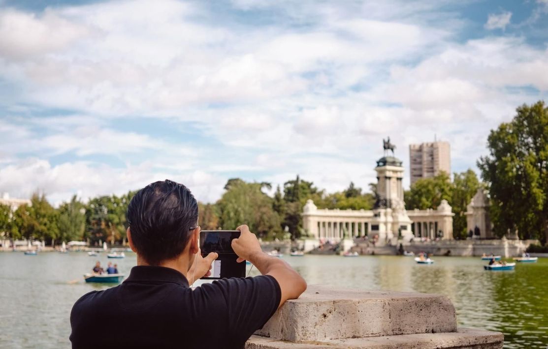 Persona fotografiando un monumento junto a un lago con una fuente y árboles bajo un cielo parcialmente nublado.