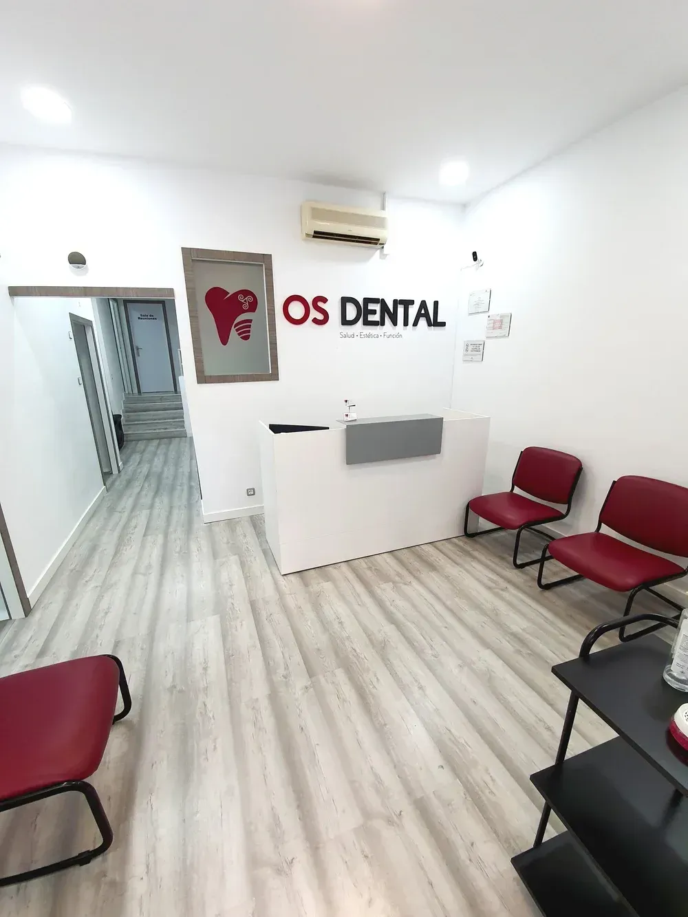 Sala de espera luminosa de la clínica dental con sillas rojas, paredes blancas y mostrador de recepción de OS Dental.