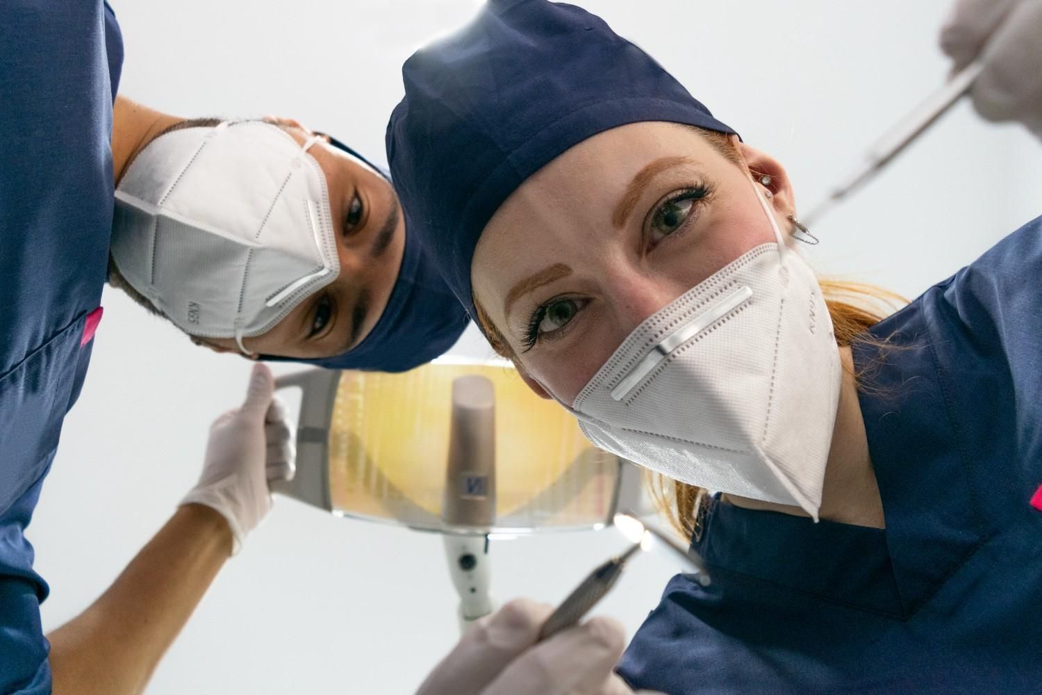 Dos miembros del personal médico enmascarados, vestidos con uniformes azules, inclinados sobre un paciente, vistos desde abajo.