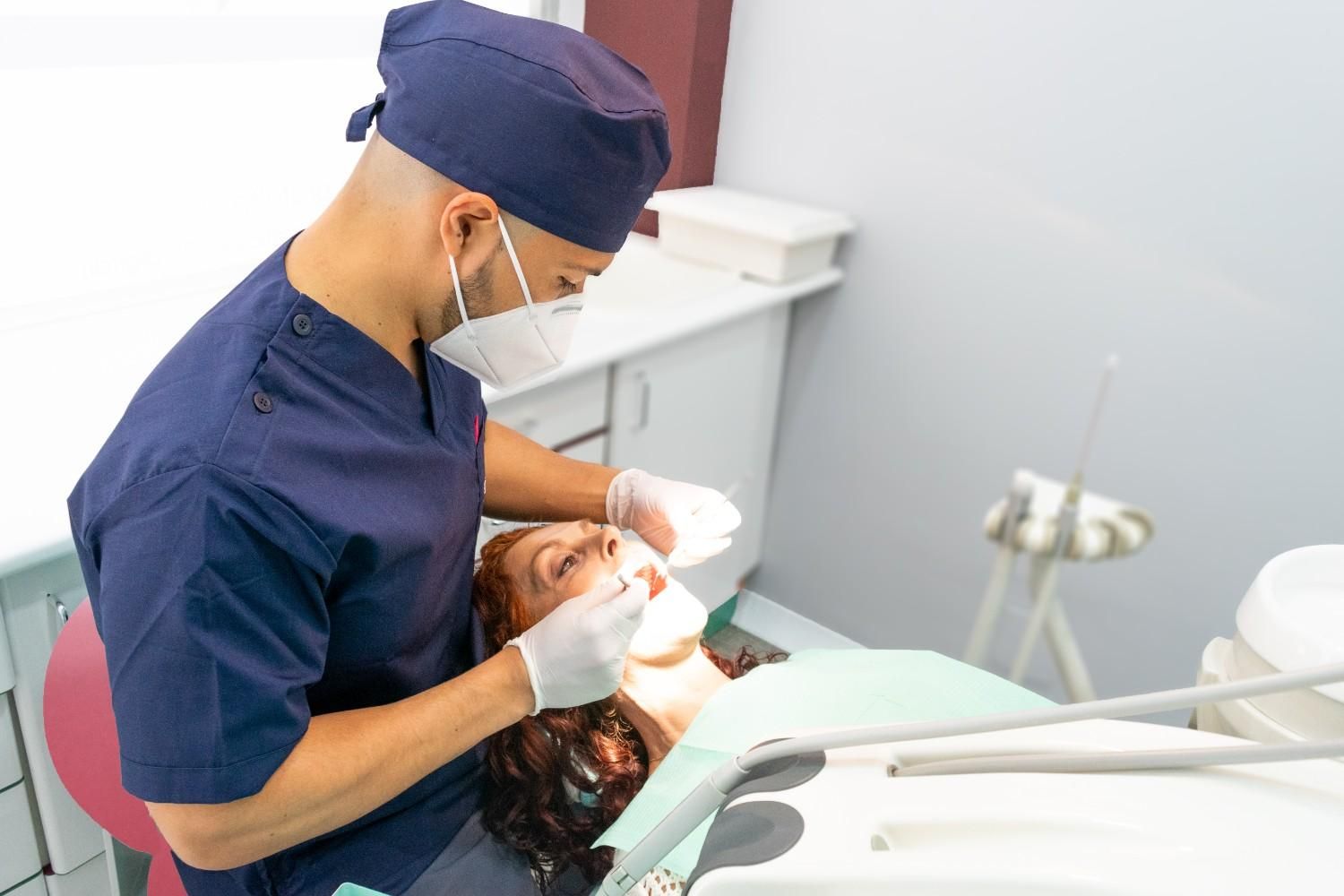 Dentist examining a patient in a dental clinic, wearing blue scrubs and a mask.