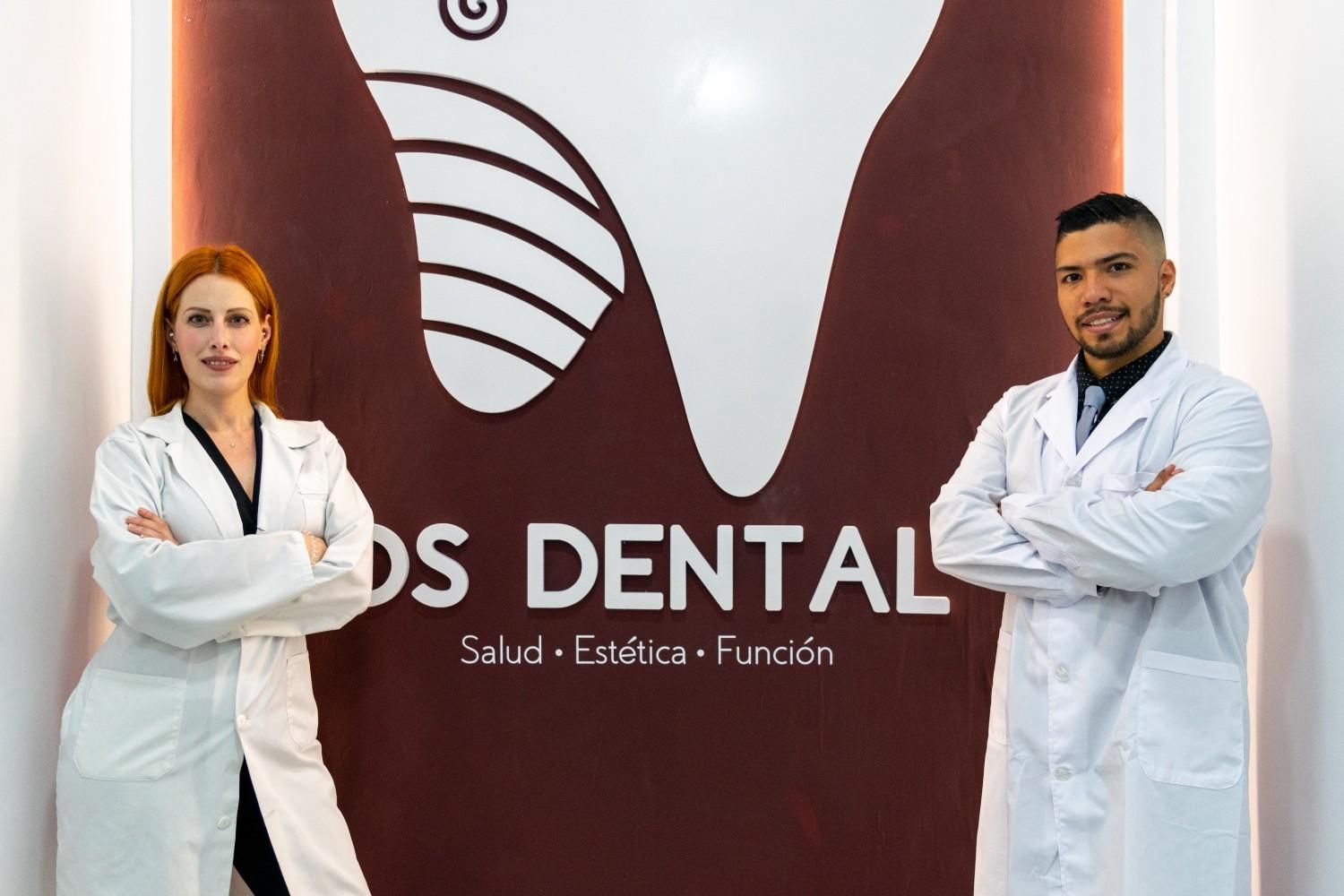 Dos dentistas con batas blancas, de pie con los brazos cruzados frente al letrero de una clínica dental.
