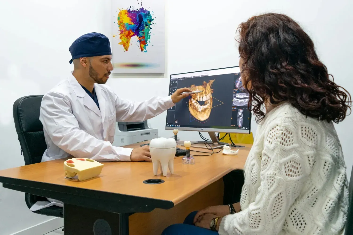 Un médico le muestra una tomografía dental a un paciente sentado en el consultorio de una clínica, con un modelo 3D del diente sobre el escritorio.