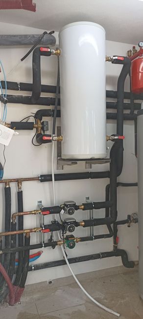 Un calentador de agua blanco está ubicado en una habitación rodeado de tuberías.