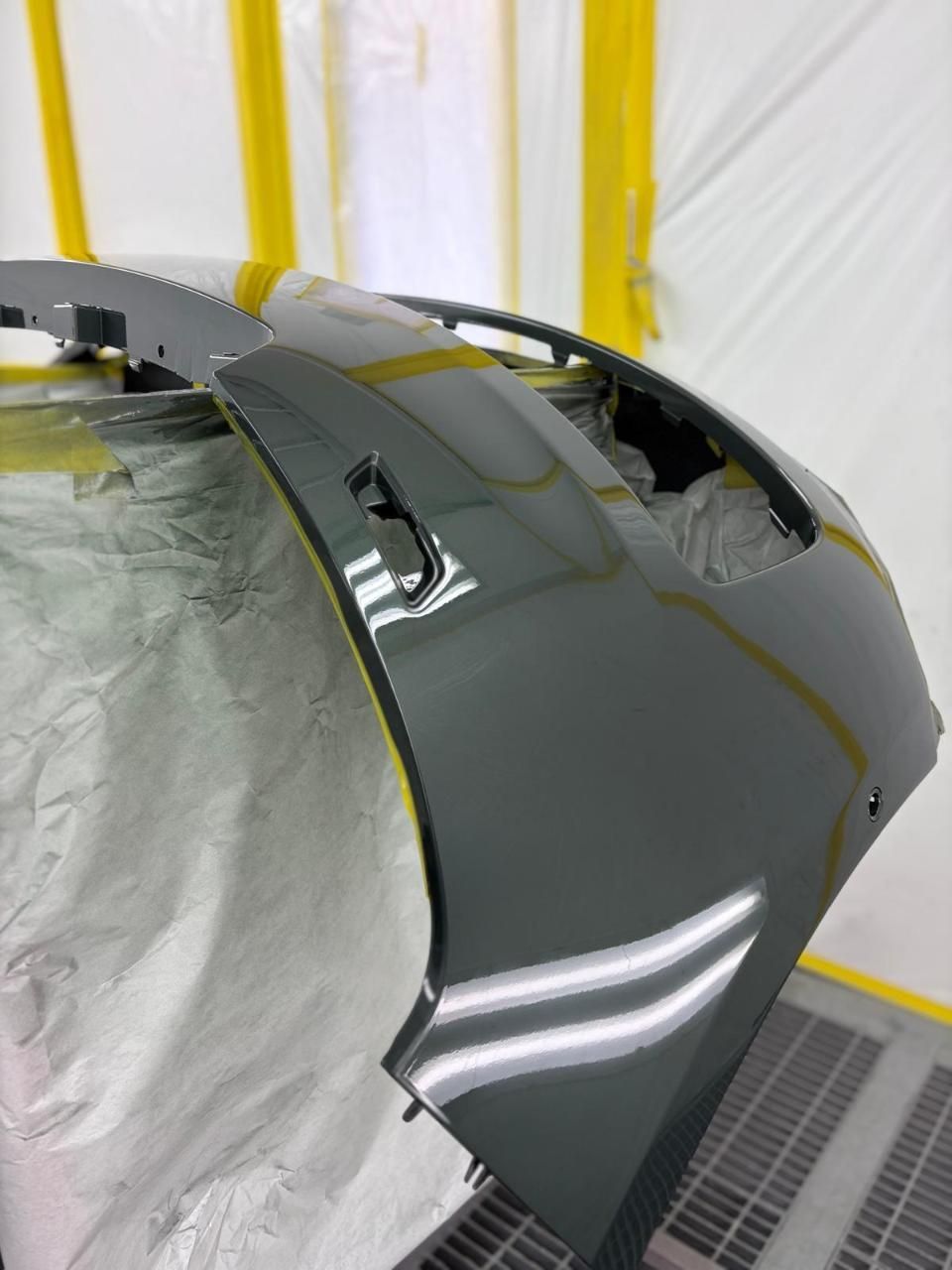 Paraurti di un'auto grigio scuro in una cabina di verniciatura a spruzzo, con nastro adesivo e teli di plastica a copertura delle aree circostanti.