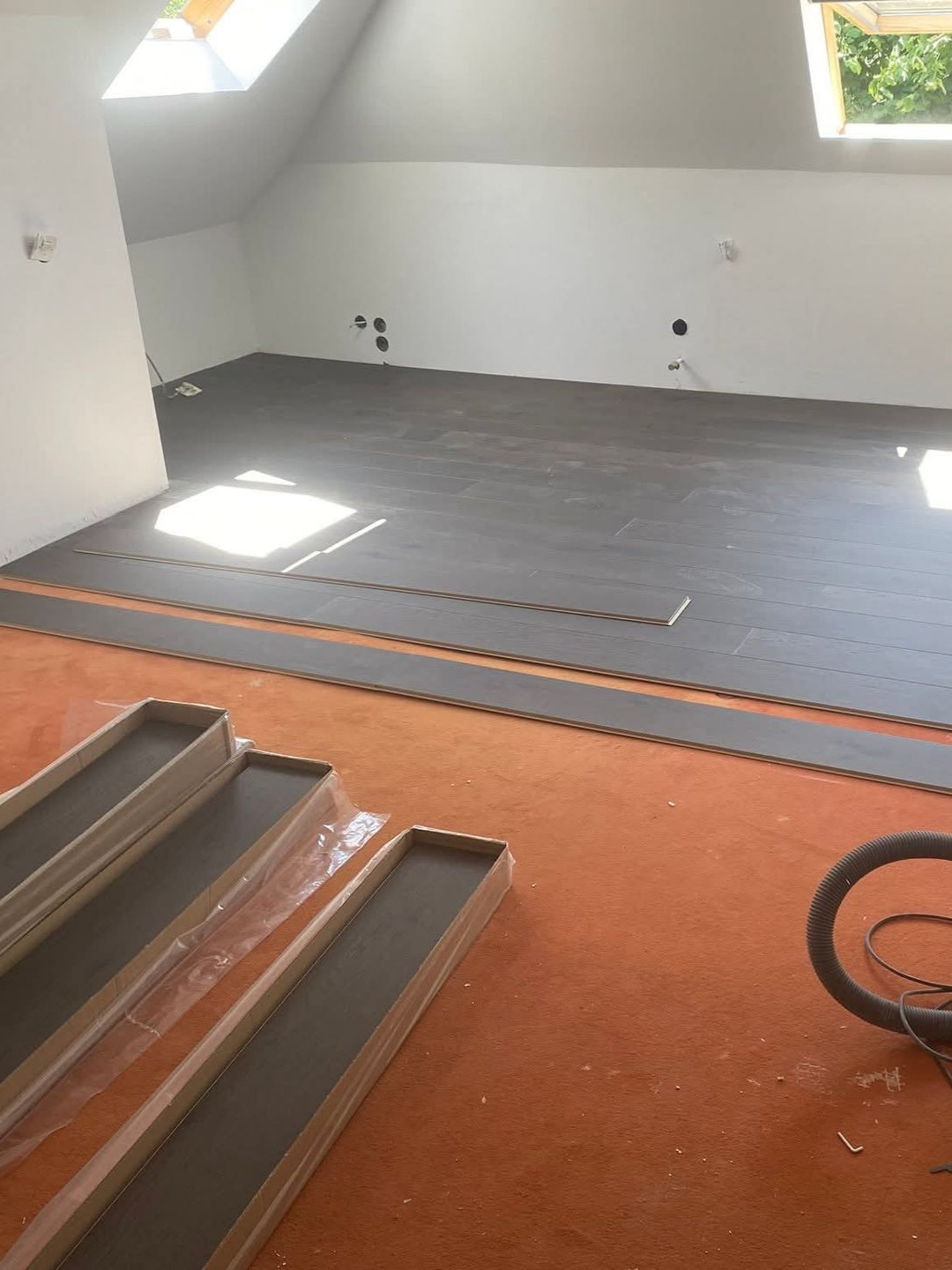 Une pièce dont le sol est partiellement posé. Le revêtement de sol noir recouvre la majeure partie de l'espace. Au premier plan, on aperçoit de la moquette marron.