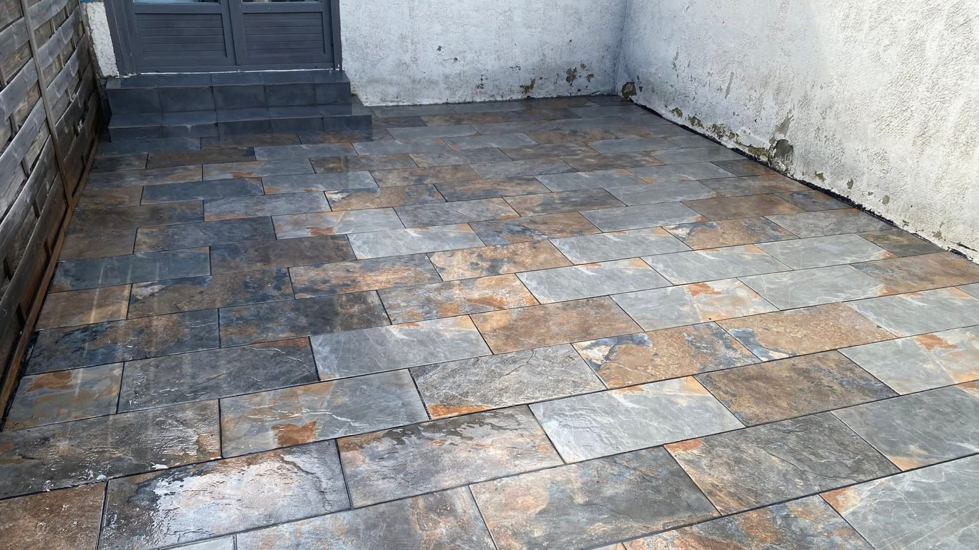 Terrasse en pierre avec des marches menant à une porte sombre, ornée de dalles en ardoise multicolores.