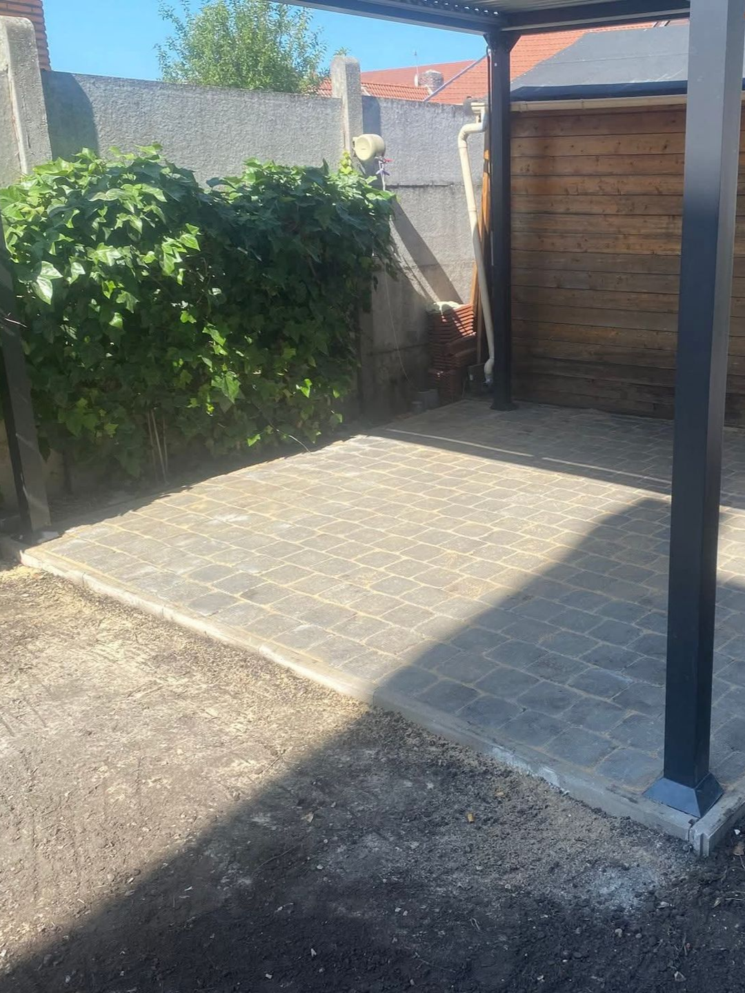 Une petite terrasse en briques sous une pergola, jouxtant une clôture en bois et des arbustes envahissants.