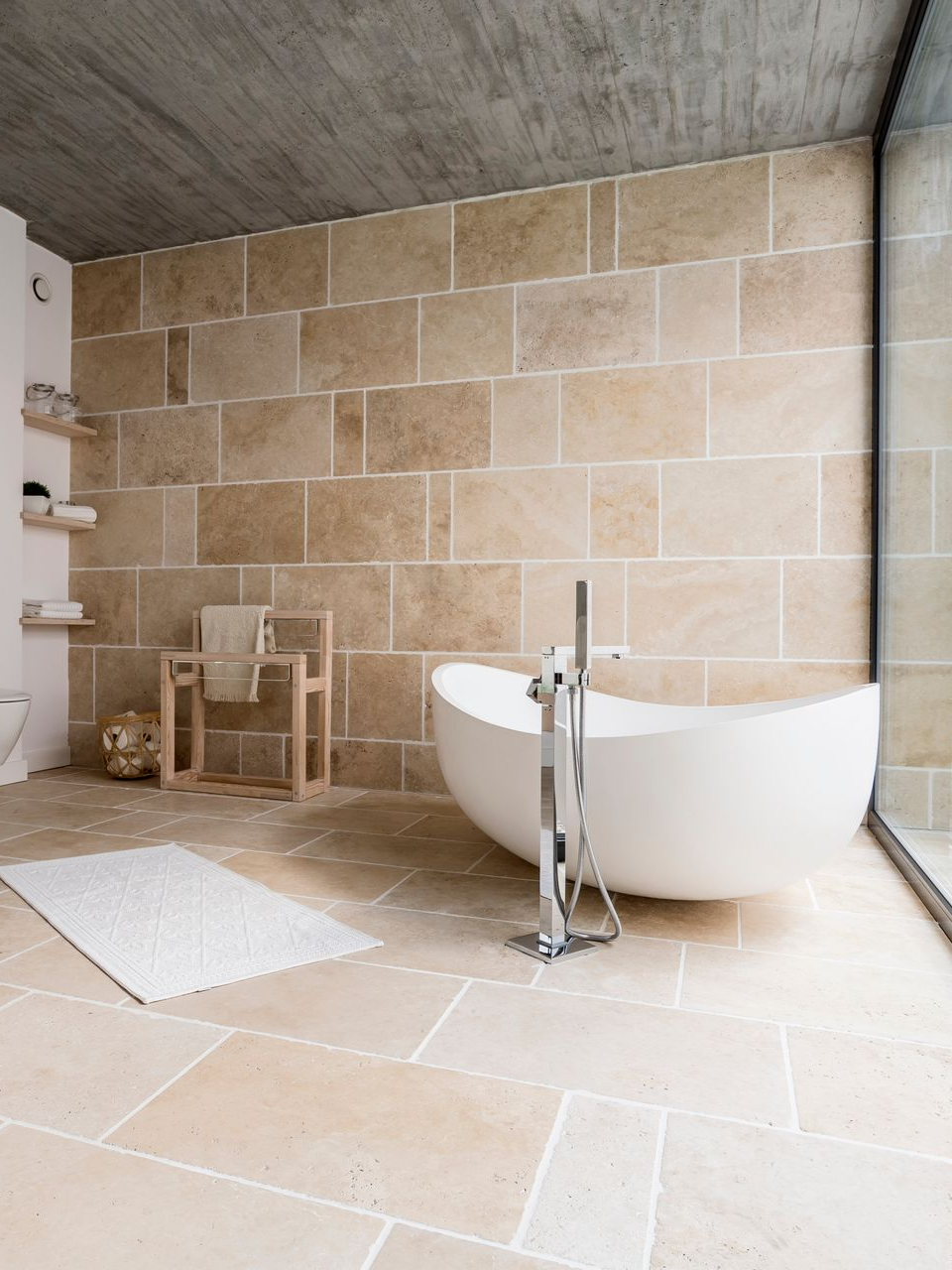 Salle de bains moderne avec murs en pierre beige, baignoire blanche et douche ouverte.