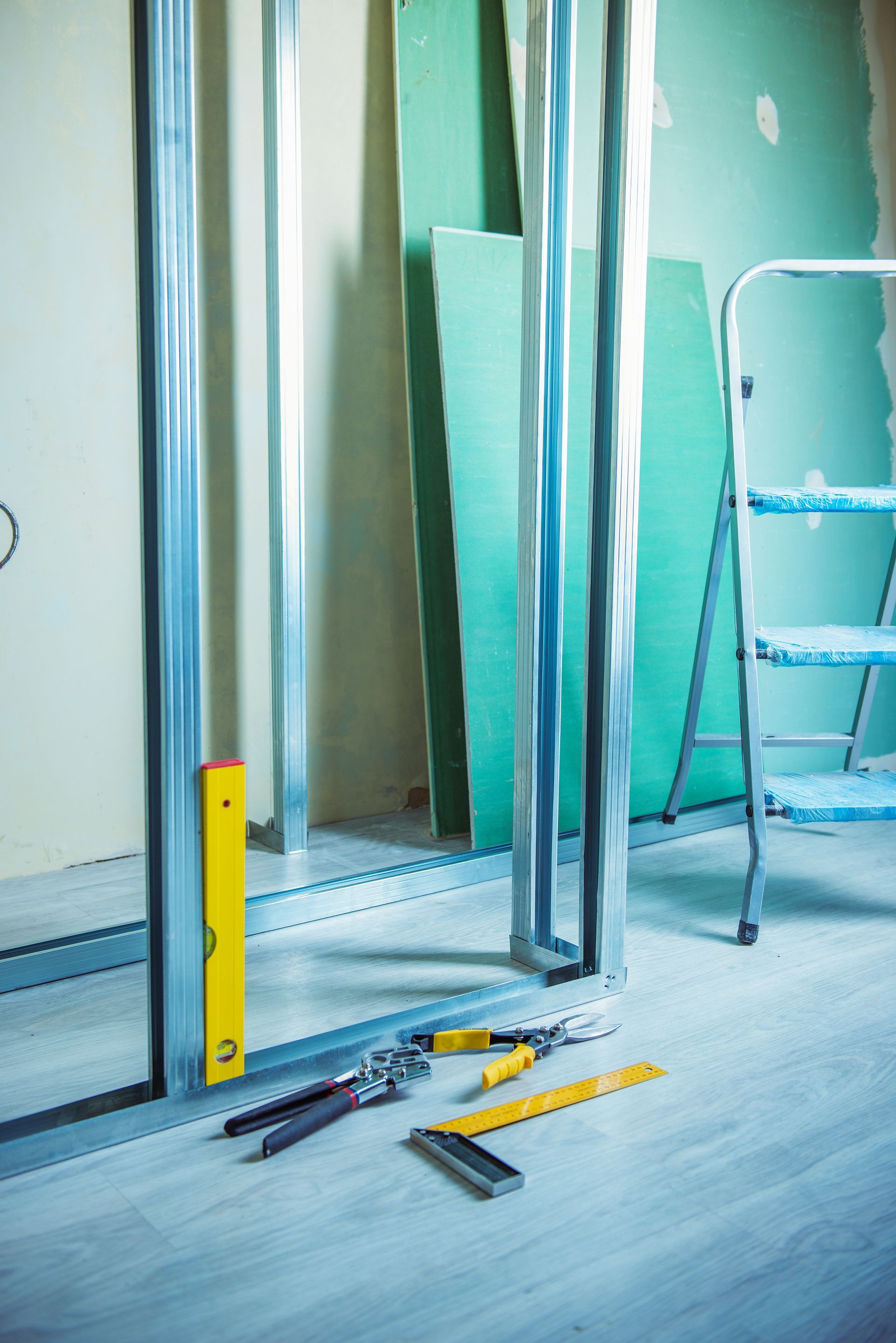 Scène de chantier : structure métallique, plaques de plâtre, outils et échelle à l’intérieur d’une pièce en rénovation.